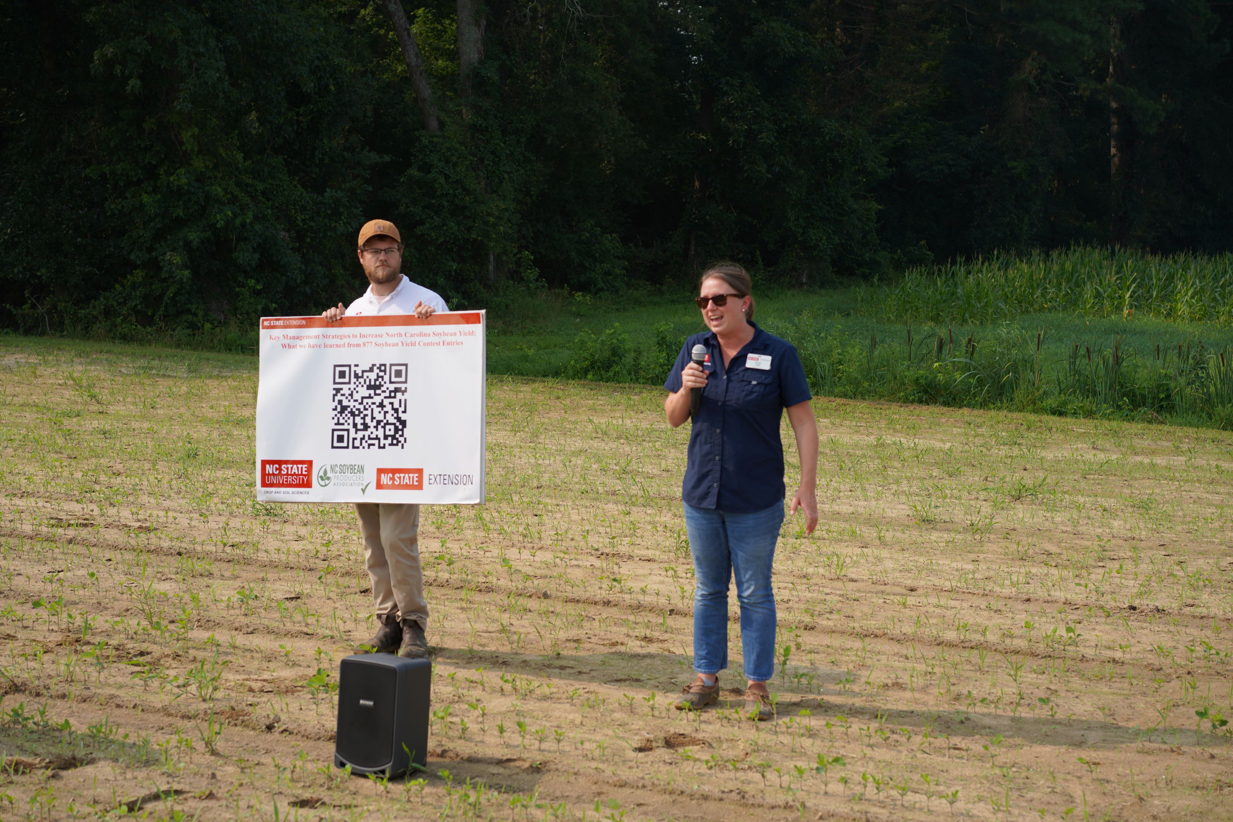 A woman gives a talk while a man holds a sign nearby.